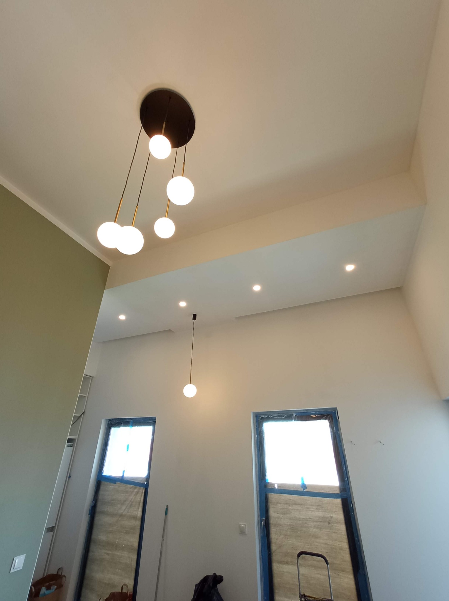 Nowoczesna lampa wisząca z kulistymi kloszami w przestronnym wnętrzu z wysokim sufitem, widoczne okna zabezpieczone folią malarską i punktowe oświetlenie sufitowe.