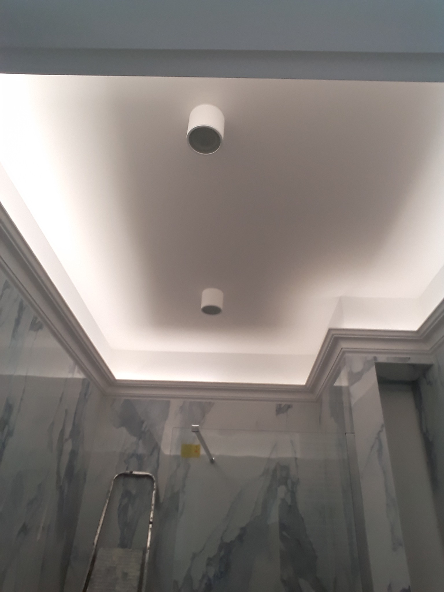 Biały sufit podwieszany z oświetleniem LED w łazience, widoczne dwa okrągłe reflektory i marmurowe ściany, drabina oparta o ścianę.