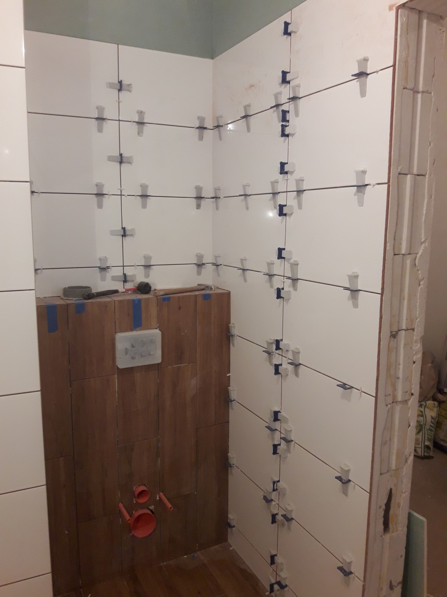 Świeżo ułożone, białe płytki ścienne w łazience, z użyciem systemu poziomowania płytek (klipsy i kliny), kontrastujące z drewnopodobnymi płytkami na ścianie pod prysznicem, widoczne narzędzia...