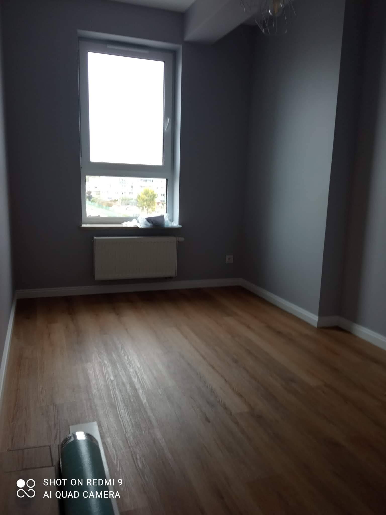 Pusty pokój w Markach z drewnianą podłogą i szarymi ścianami po świeżym malowaniu, widoczne okno z grzejnikiem i rolka folii na podłodze.