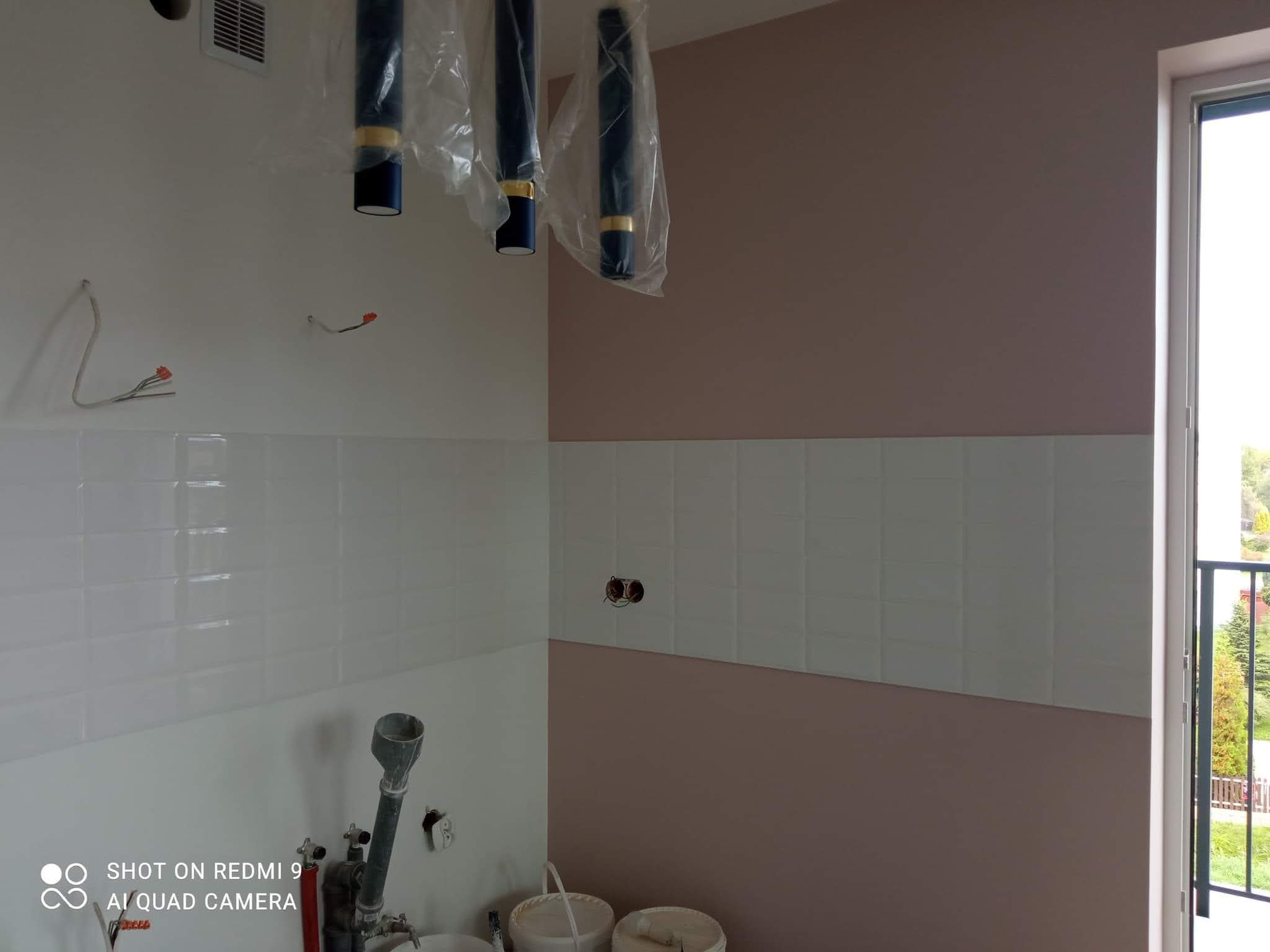 Świeżo pomalowane ściany w pomieszczeniu, częściowo wyłożone białymi, prostokątnymi płytkami. Widoczne puszki elektryczne i wiszące, zabezpieczone folią lampy. Na podłodze stoją wiadra z farbą...