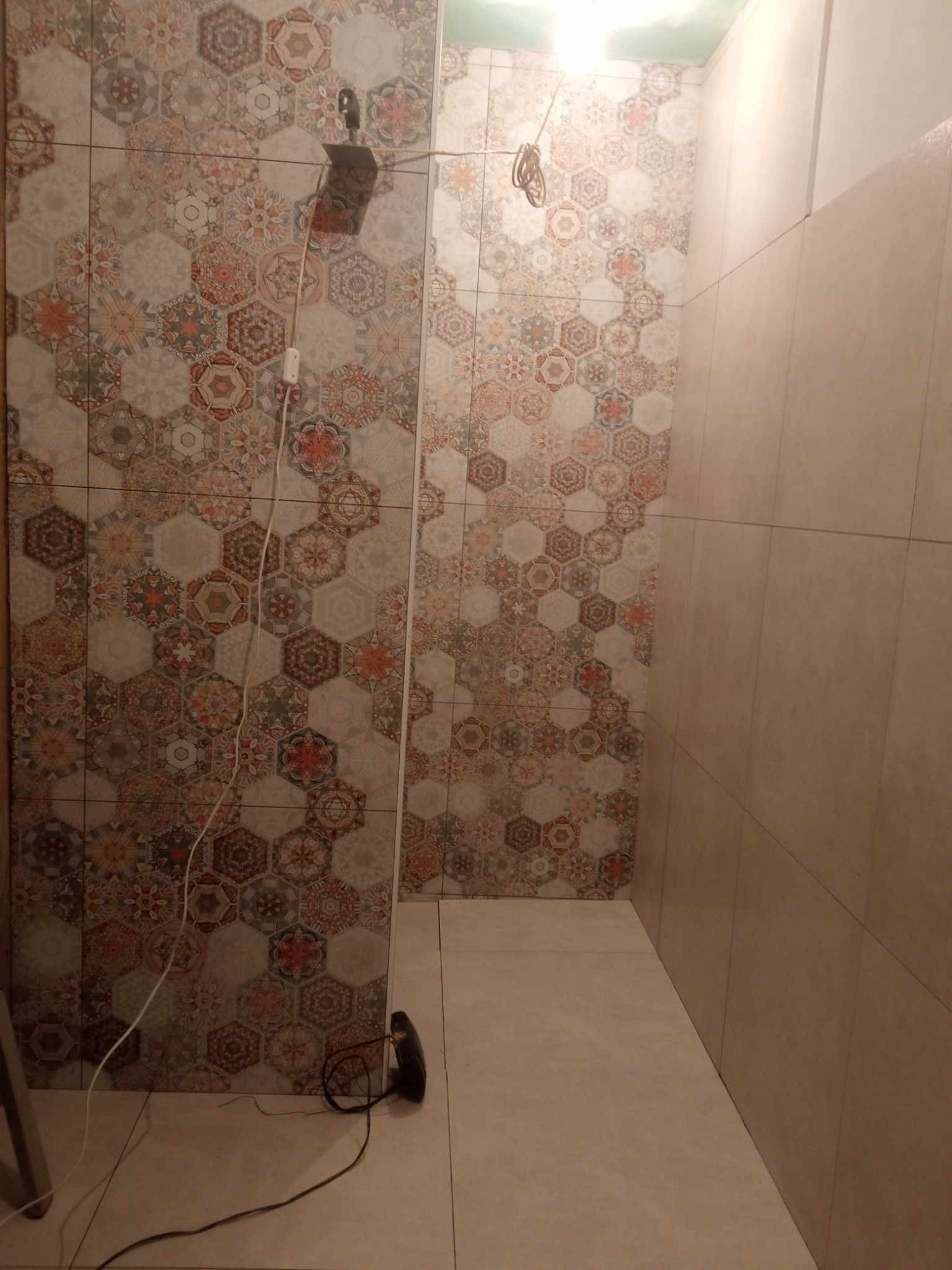 Fragment łazienki w trakcie remontu, ściana z heksagonalnymi płytkami o wzorze patchworkowym kontrastuje z gładkimi, beżowymi płytkami po prawej stronie, widoczne narzędzia i przewody elektryczne.