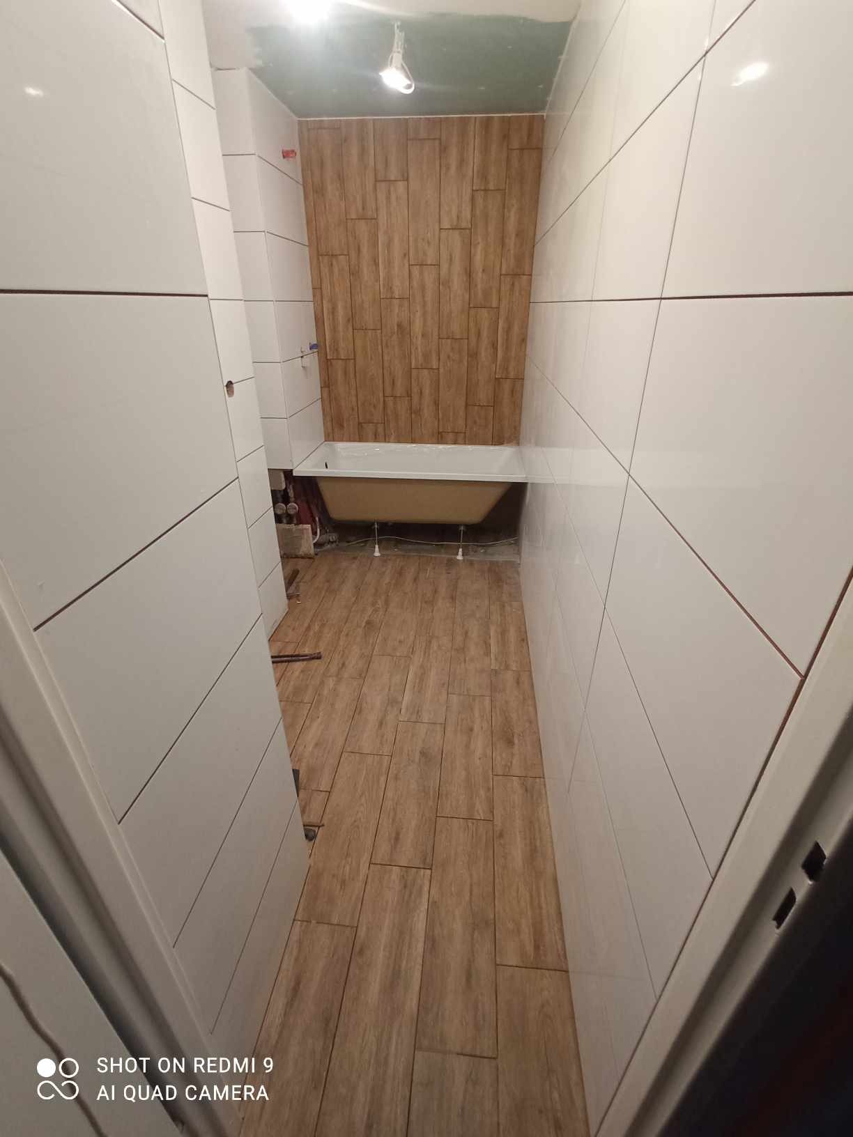 Wąska łazienka w trakcie remontu: białe płytki na ścianach kontrastują z drewnopodobną podłogą i ścianą za wanną, widoczna wanna na nóżkach i niedokończone prace na suficie.