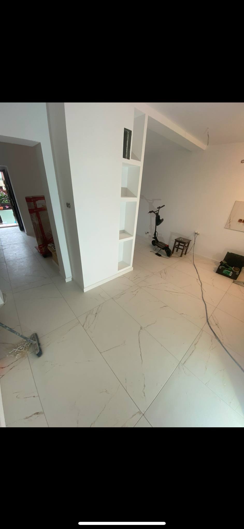 Białe, marmurowe płytki podłogowe w jasnym wnętrzu po remoncie. Widoczny mop i narzędzia. Podział pomieszczenia ścianką z półkami.