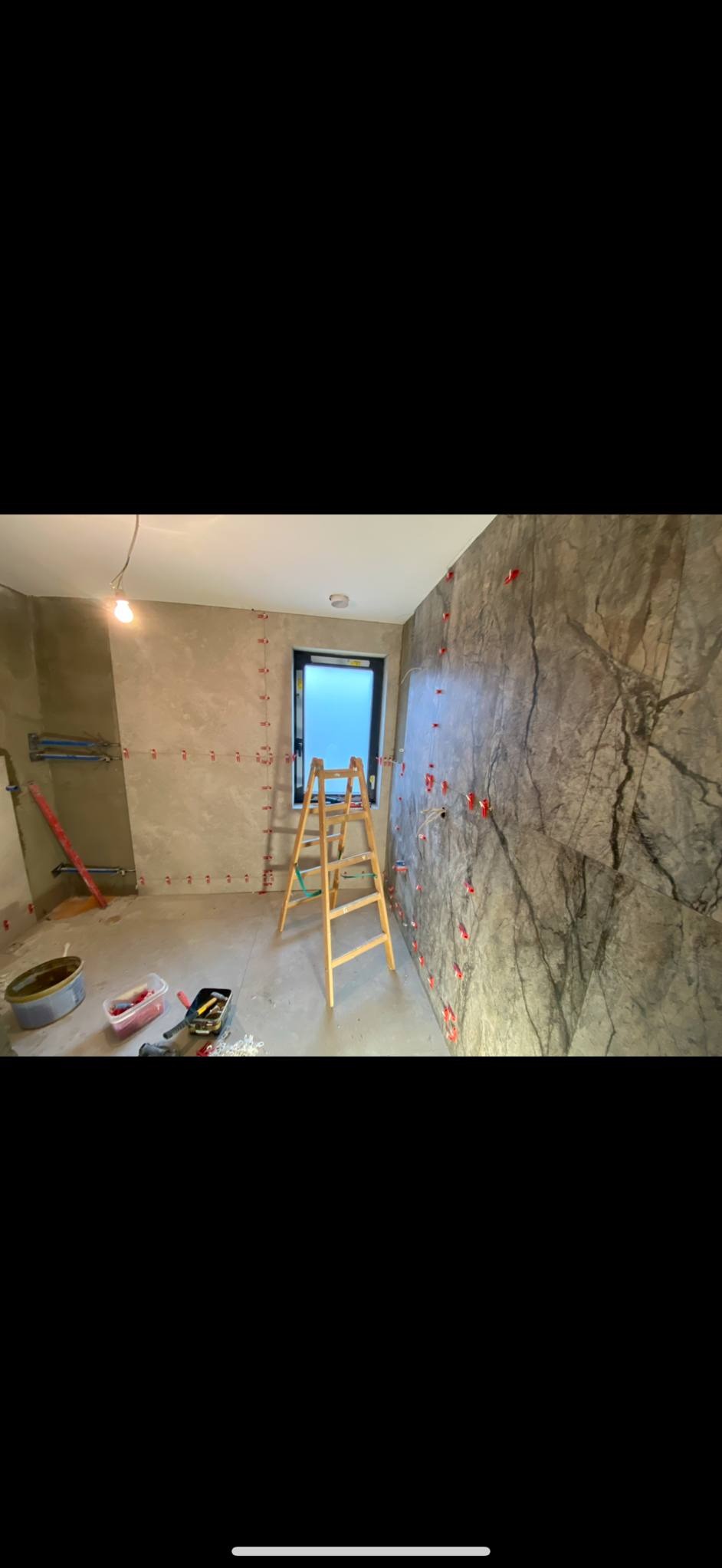 Wnętrze łazienki w trakcie prac wykończeniowych. Na ścianach marmuropodobne płytki z czerwonymi krzyżykami dystansowymi. Na podłodze drabina i narzędzia.