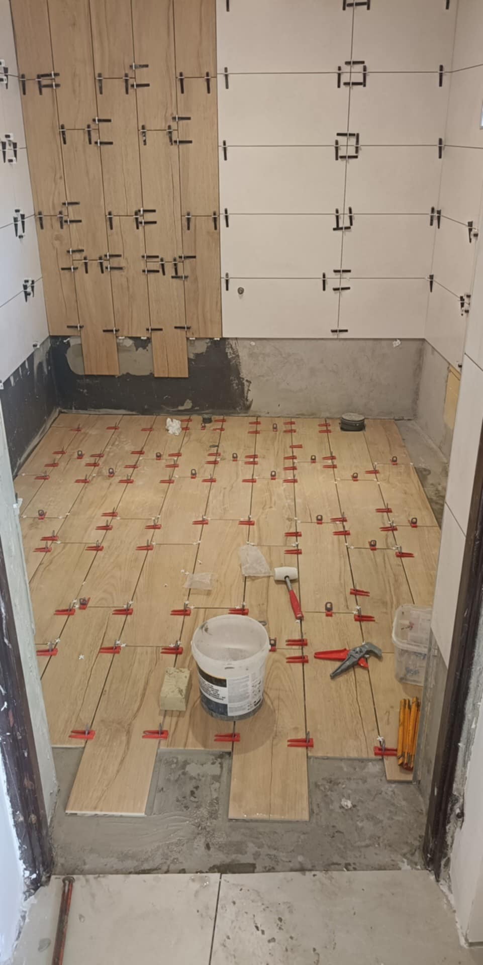 Układanie płytek podłogowych imitujących drewno z użyciem systemu poziomowania. Widoczne krzyżyki dystansowe, młotek i wiadro z zaprawą. Prace w toku.