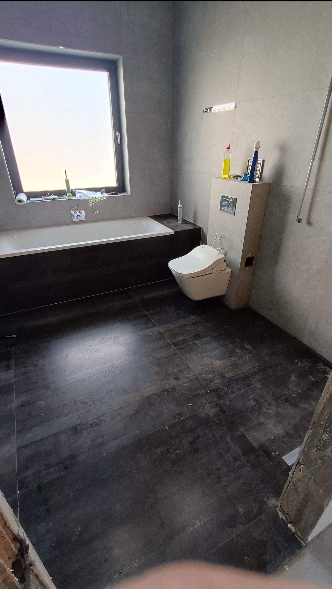 Nowoczesna łazienka z ciemną podłogą i szarymi płytkami. Widoczna wanna, toaleta myjąca i okno. Na ścianie narzędzia i chemia budowlana.