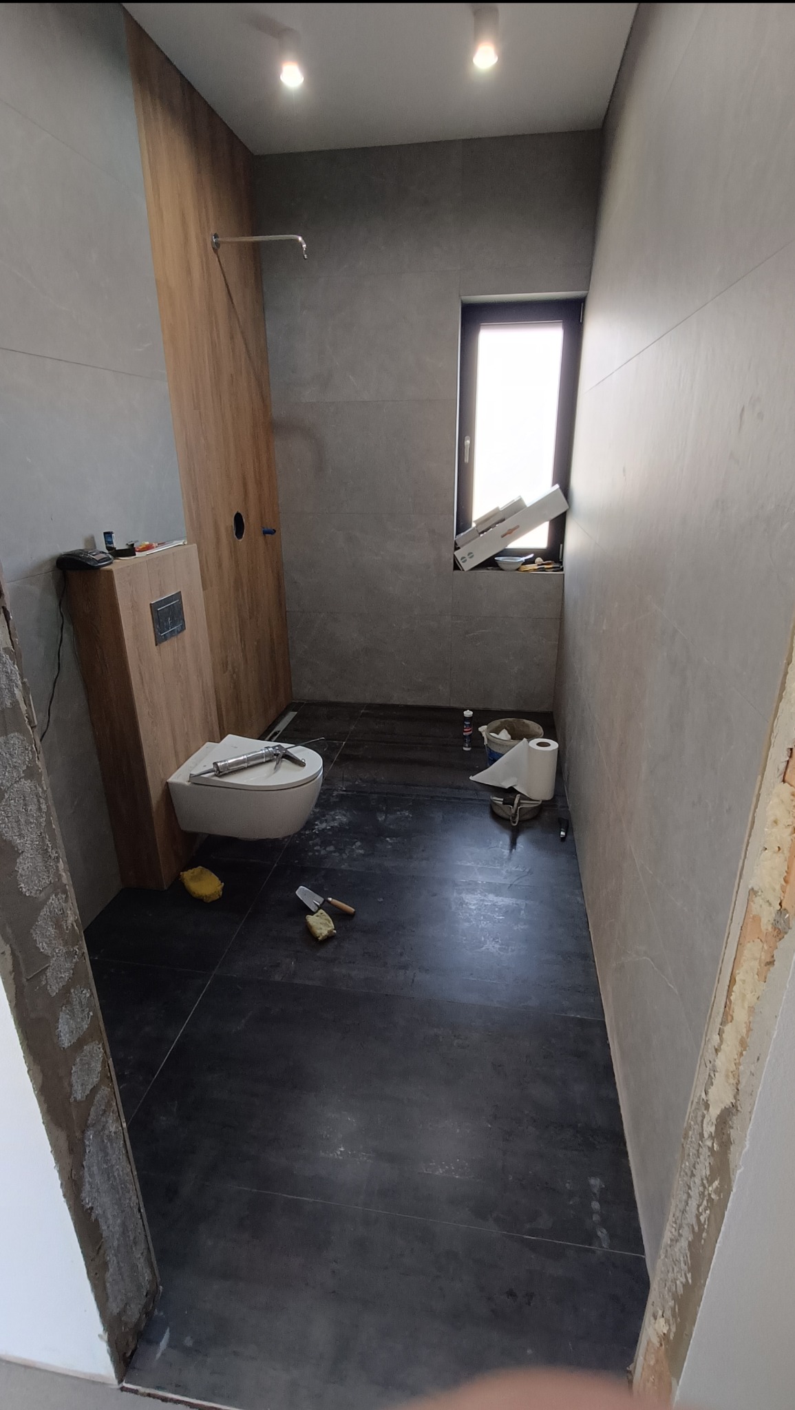 Montaż płytek w łazience z podwieszaną toaletą, widoczne narzędzia i resztki materiałów budowlanych. Wykończenie ściany imitujące drewno.