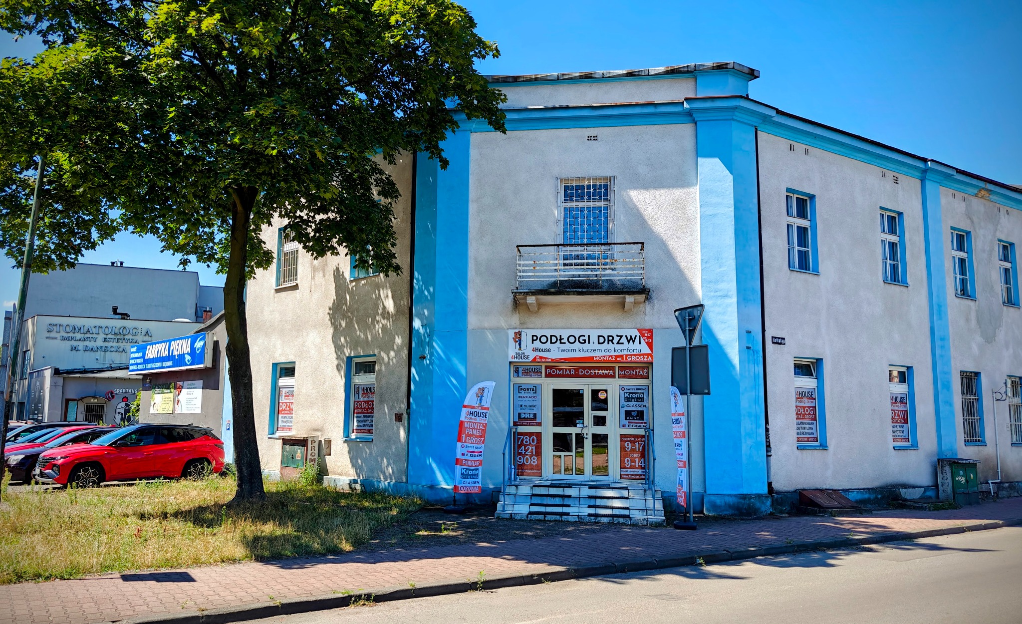 Fasada budynku z reklamami firmy oferującej podłogi i drzwi, widoczne banery reklamowe, okna z plakatami informacyjnymi oraz wejście do sklepu z ekspozycją.