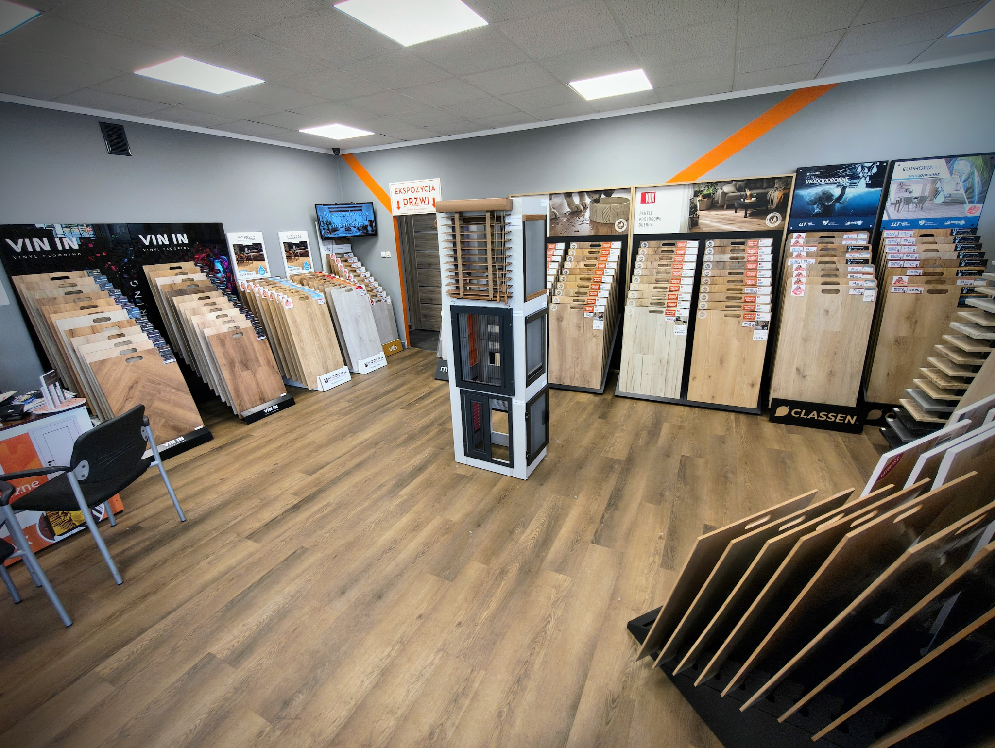 Salon z ekspozycją paneli podłogowych różnych marek i wzorów, prezentowanych na stojakach i ścianach, w jasnym wnętrzu z oświetleniem sufitowym.