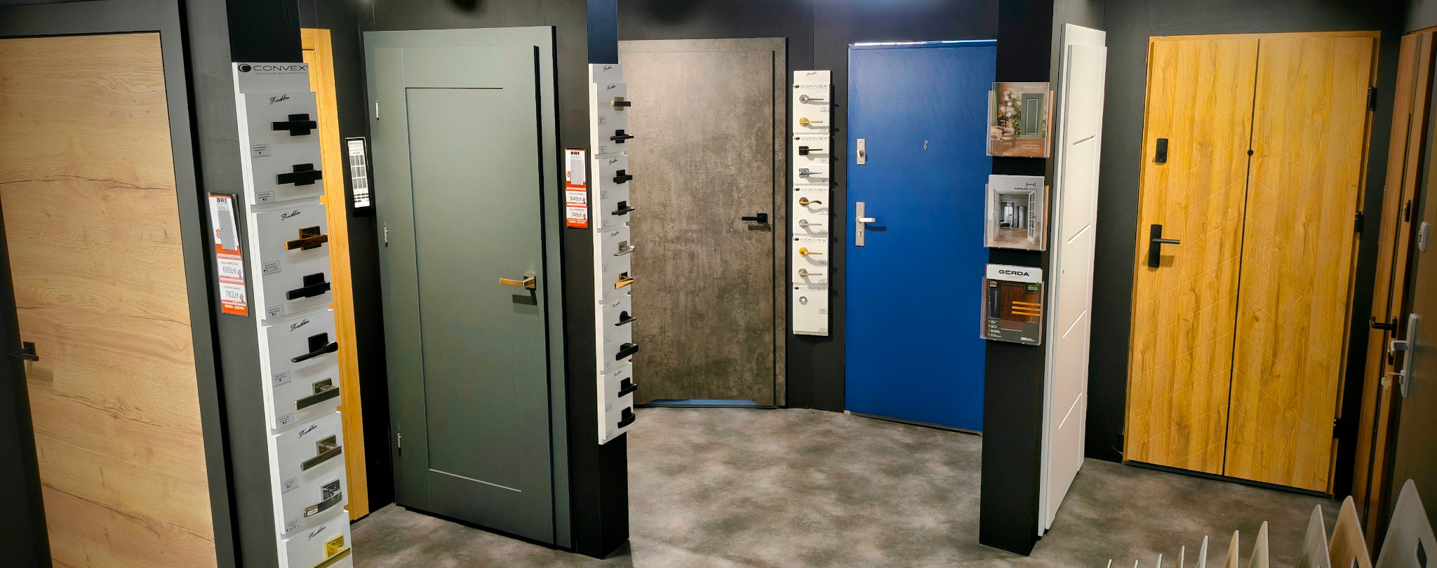 Ekspozycja różnorodnych drzwi wewnętrznych i zewnętrznych w salonie, z katalogami klamek i okuć, ukazująca szeroki wybór wzorów i kolorów.