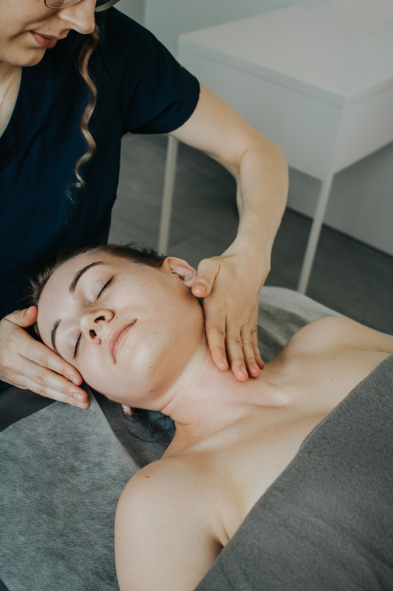 Kobieta z zamkniętymi oczami leży na szarym prześcieradle podczas delikatnego zabiegu na szyi i dekolcie wykonywanego przez masażystkę w okularach.