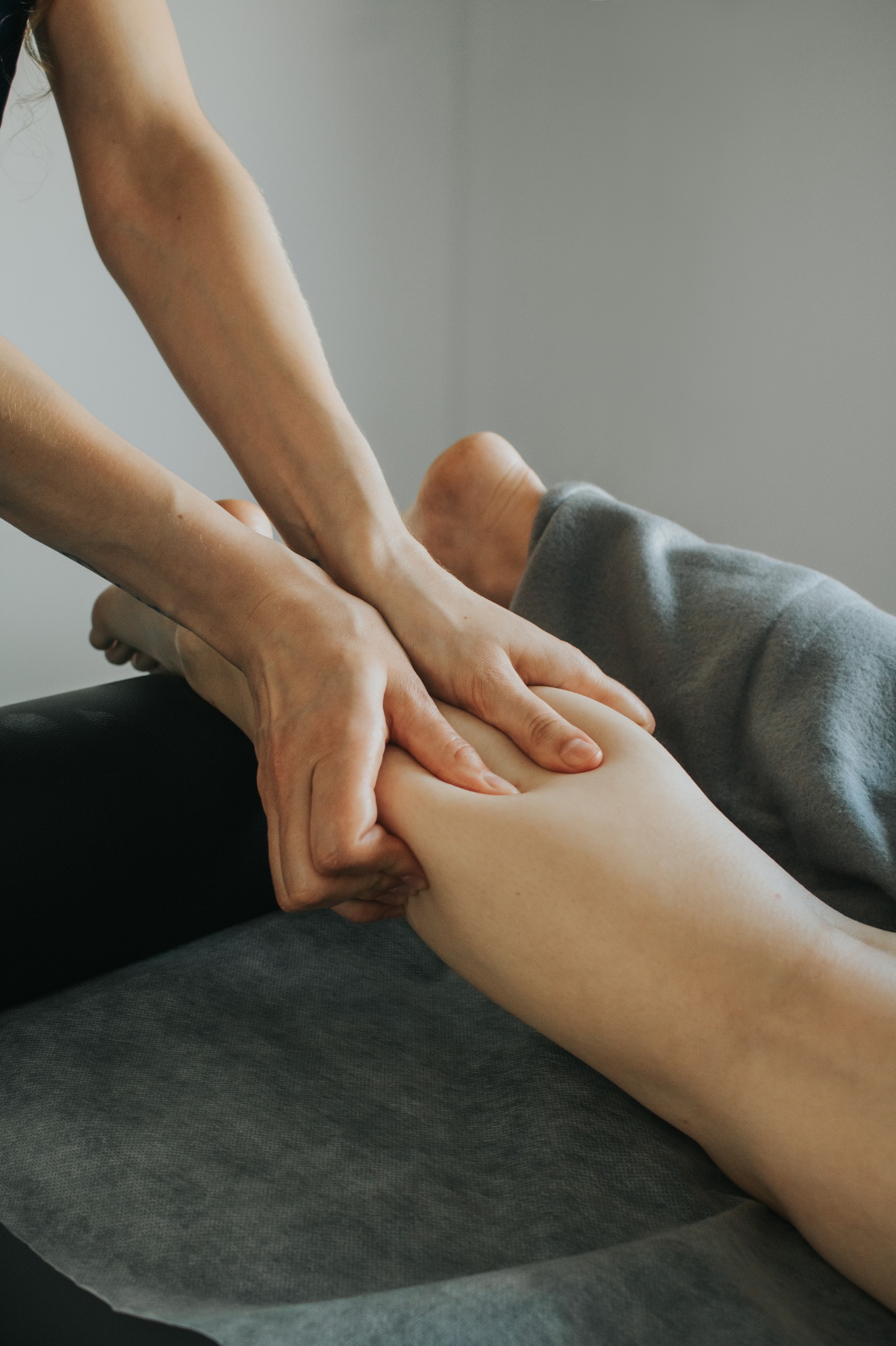 Terapeuta uciska dłońmi łydkę klienta leżącego na stole przykrytym szarą tkaniną.