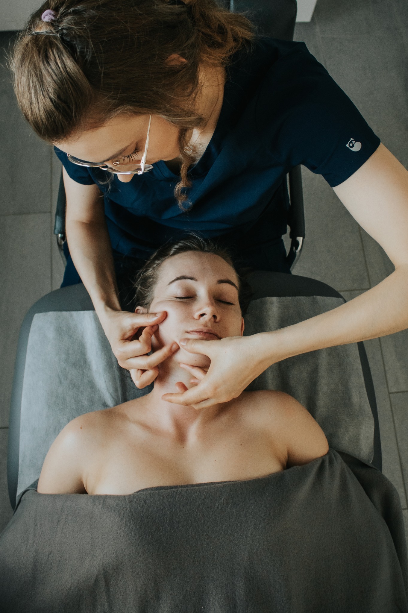 Masaż twarzy wykonywany przez masażystkę w okularach, klientka leży na szarym ręczniku na łóżku do masażu.