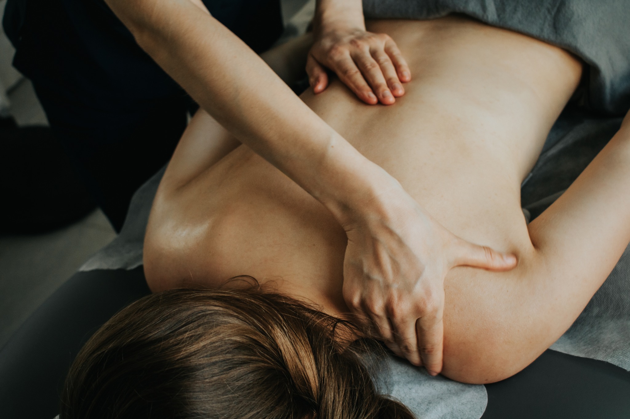 Kobieta podczas zabiegu na plecach, widoczne dłonie terapeuty uciskające mięśnie, głowa pacjentki z brązowymi włosami spoczywa na stole.