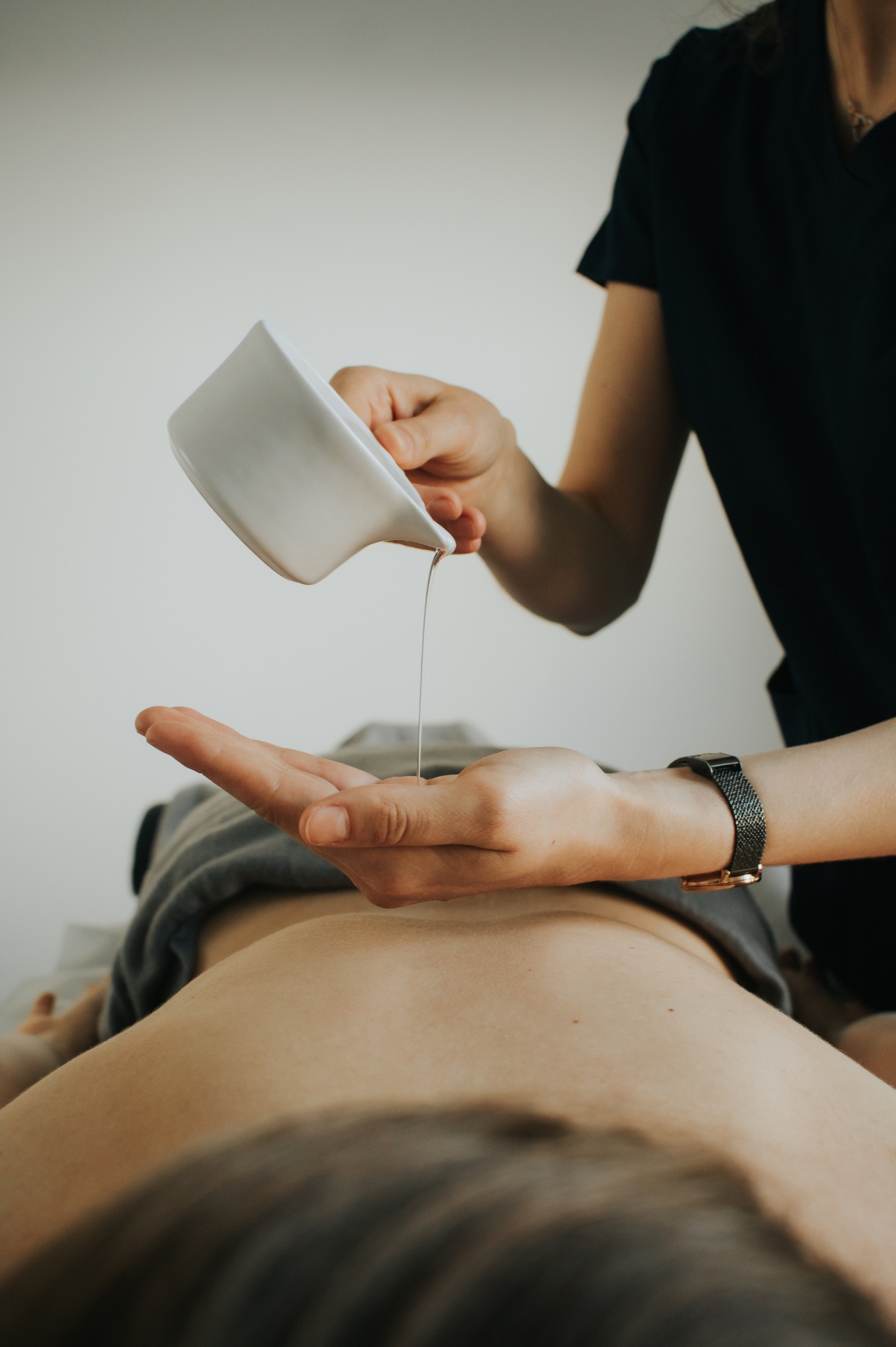 Olej wylewany z białego dzbanka na dłoń klienta leżącego na stole do zabiegów, widoczny fragment zegarka na nadgarstku terapeuty.