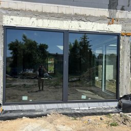 LUXFENSTER SPÓŁKA Z OGRANICZONĄ ODPOWIEDZIALNOŚCIĄ - Nowo zamontowane, duże okno aluminiowe z widocznym odbiciem osoby robiącej zdjęcie, osadzone w surowej ścianie z betonu komórkowego.