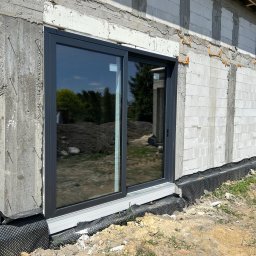 LUXFENSTER SPÓŁKA Z OGRANICZONĄ ODPOWIEDZIALNOŚCIĄ - Montaż przesuwnych, szarych drzwi okiennych w surowym stanie budynku z betonu komórkowego, widoczna folia kubełkowa przy fundamencie.