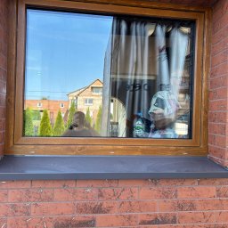 LUXFENSTER SPÓŁKA Z OGRANICZONĄ ODPOWIEDZIALNOŚCIĄ - Zamontowane okno z brązową ramą w murze z czerwonej cegły, widoczne odbicie osoby robiącej zdjęcie i częściowo zasłonięte wnętrze folią ochronną i zasłonami.