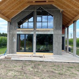 LUXFENSTER SPÓŁKA Z OGRANICZONĄ ODPOWIEDZIALNOŚCIĄ - Surowy stan domu w trakcie budowy z zamontowanymi oknami o czarnych ramach, widoczne drewniane elementy konstrukcji dachu i surowe betonowe ściany.