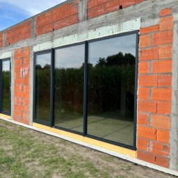 LUXFENSTER SPÓŁKA Z OGRANICZONĄ ODPOWIEDZIALNOŚCIĄ - Czarne okna w surowym stanie budynku z czerwonej cegły, widoczne od zewnątrz na tle zieleni i nieba. Budynek w trakcie budowy, z widocznymi elementami konstrukcyjnymi i izolacyjnymi.