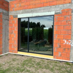 LUXFENSTER SPÓŁKA Z OGRANICZONĄ ODPOWIEDZIALNOŚCIĄ - Montaż okna aluminiowego w surowym murze z czerwonej cegły, widoczne narzędzia i folia zabezpieczająca wewnątrz budynku, pole kukurydzy w odbiciu szyby.