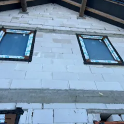 Montaż okien na poddaszu w Bytomiu, widoczne dwa okna z brązowymi ramami, zabezpieczone taśmą montażową z logo VEKA, w trakcie murowania ściany z białych bloczków.