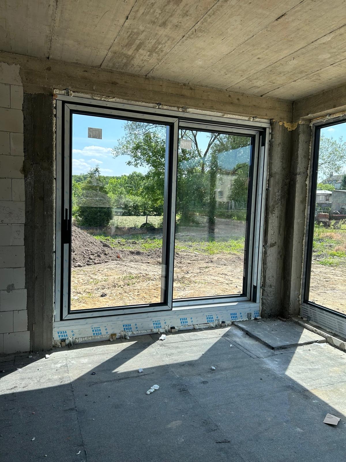 Montaż dużych przesuwnych okien z czarnymi ramami w surowym stanie, widok na zewnątrz z zielenią i fragmentem budynku w tle, pianka montażowa widoczna u dołu ramy.