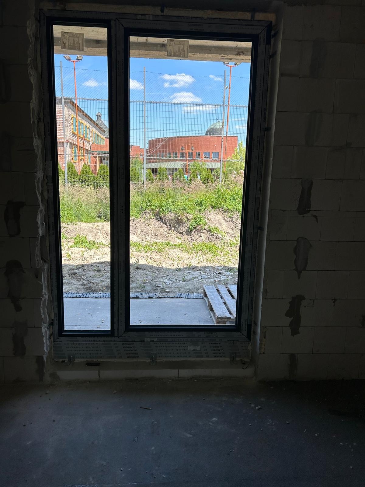 Nowe okno z czarną ramą, widok na plac budowy z budynkami w tle, surowy stan wykończenia wnętrza.