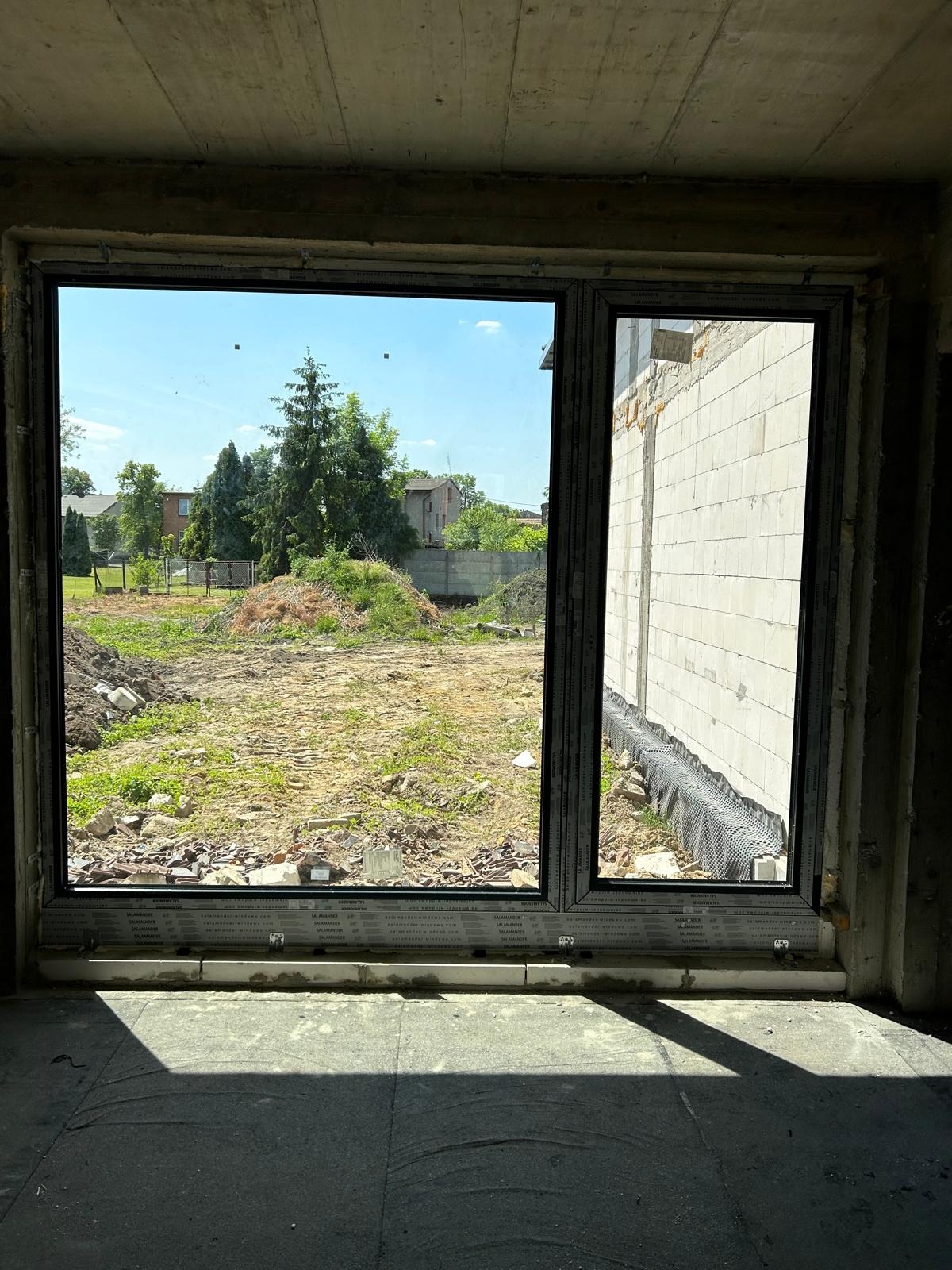 Nowe dwudzielne okno z czarnymi ramami, widok na niezagospodarowany teren budowy i fragment ściany z pustaków.