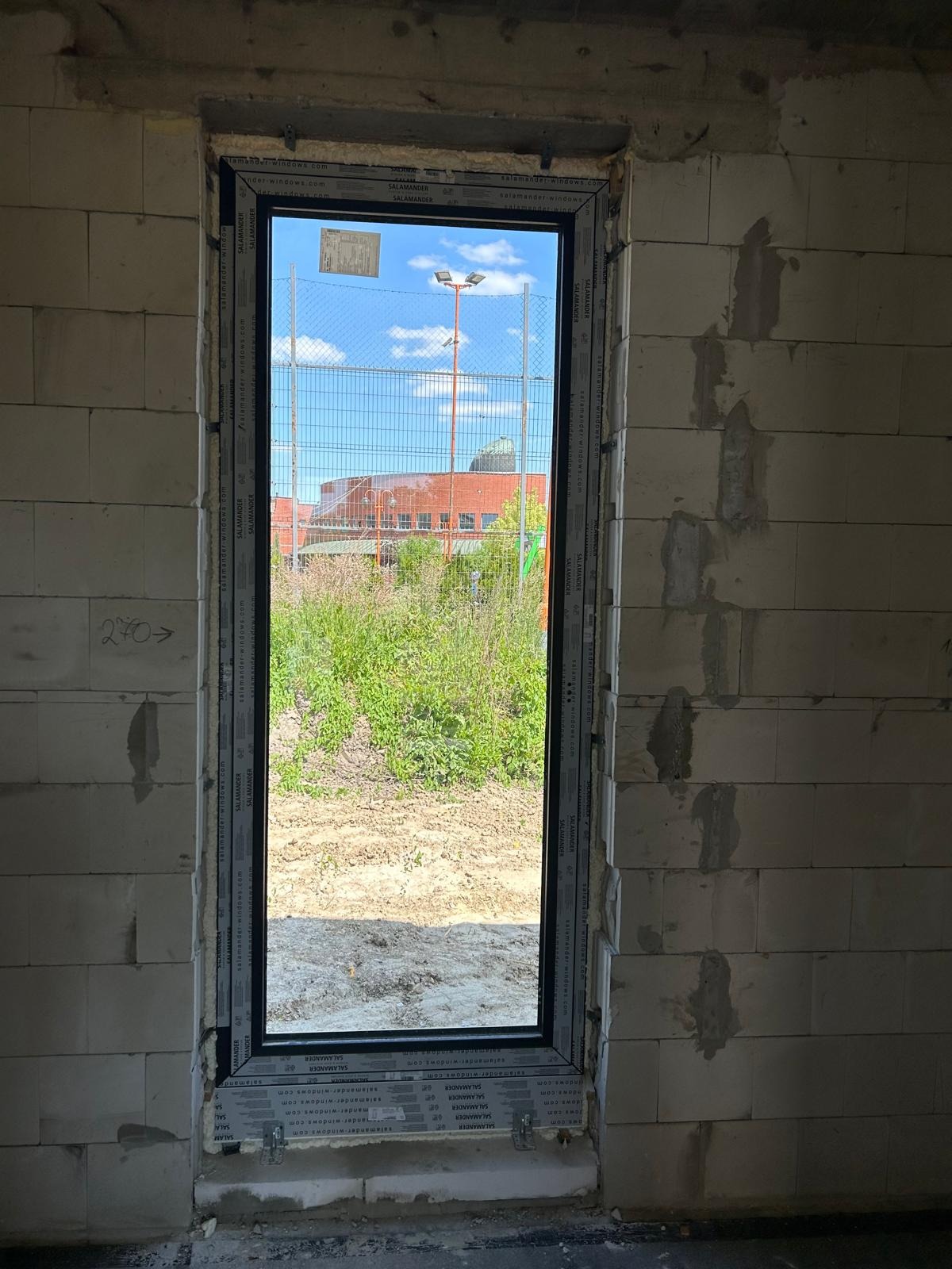 Nowo zamontowane okno PCV w surowym stanie, widoczne ślady po zaprawie na ścianach z betonu komórkowego, na zewnątrz widok na plac z zielenią i budynek w oddali.