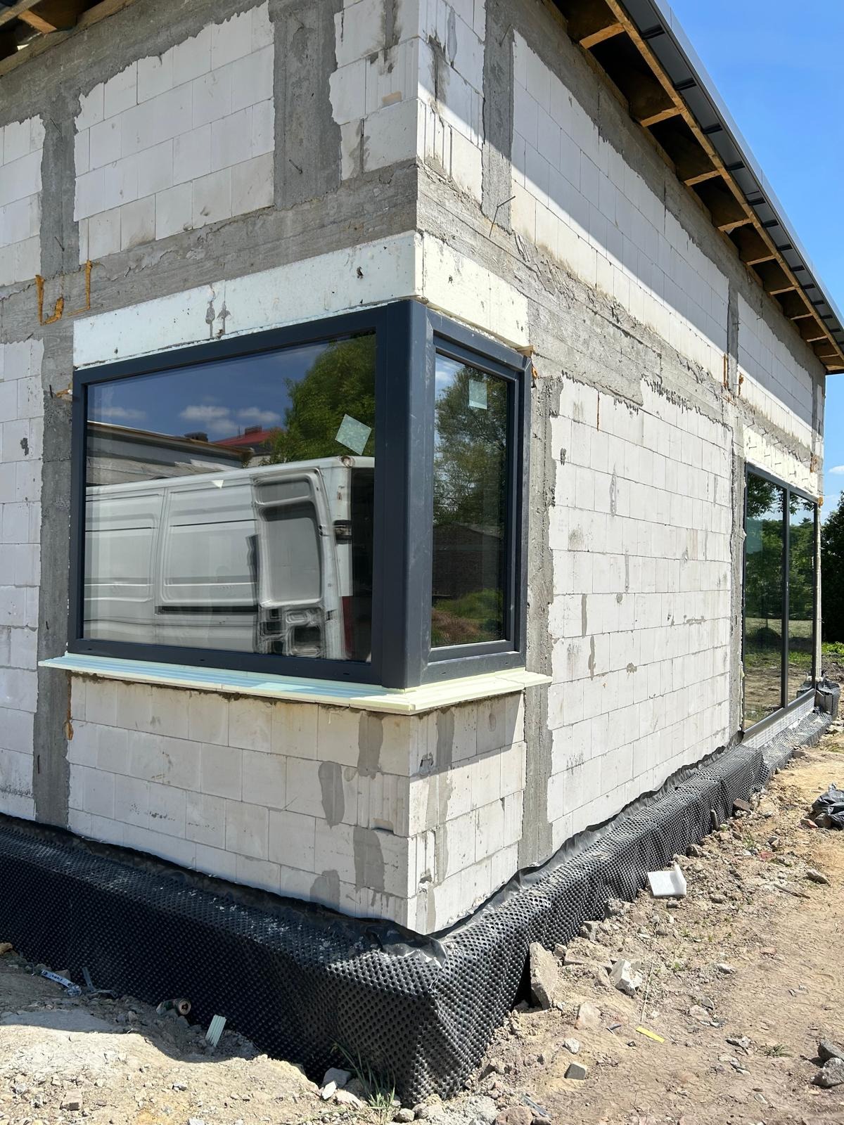 Surowy stan budynku w trakcie montażu stolarki okiennej z czarnymi ramami; widoczna biała ściana z betonu komórkowego, ocieplenie pod oknem oraz folia kubełkowa przy fundamencie, w szybie odbija...