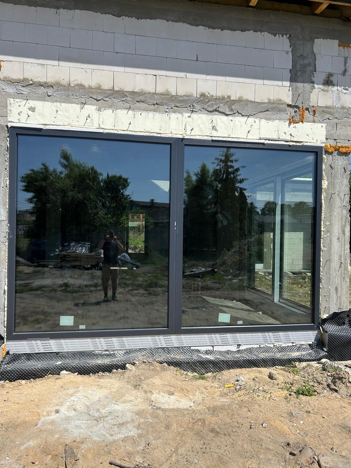 Nowo zamontowane, duże okno aluminiowe z widocznym odbiciem osoby robiącej zdjęcie, osadzone w surowej ścianie z betonu komórkowego.