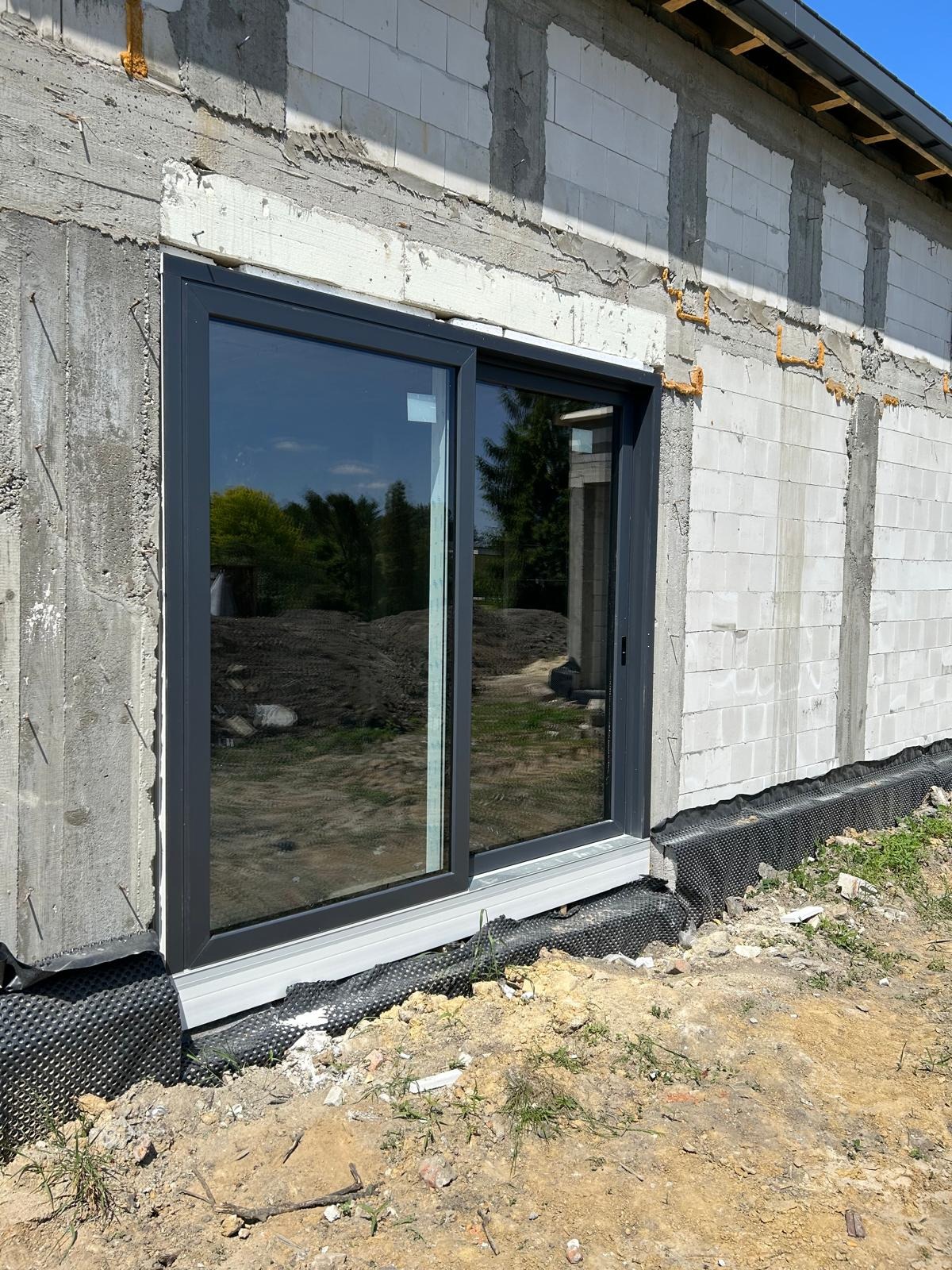 Montaż przesuwnych, szarych drzwi okiennych w surowym stanie budynku z betonu komórkowego, widoczna folia kubełkowa przy fundamencie.