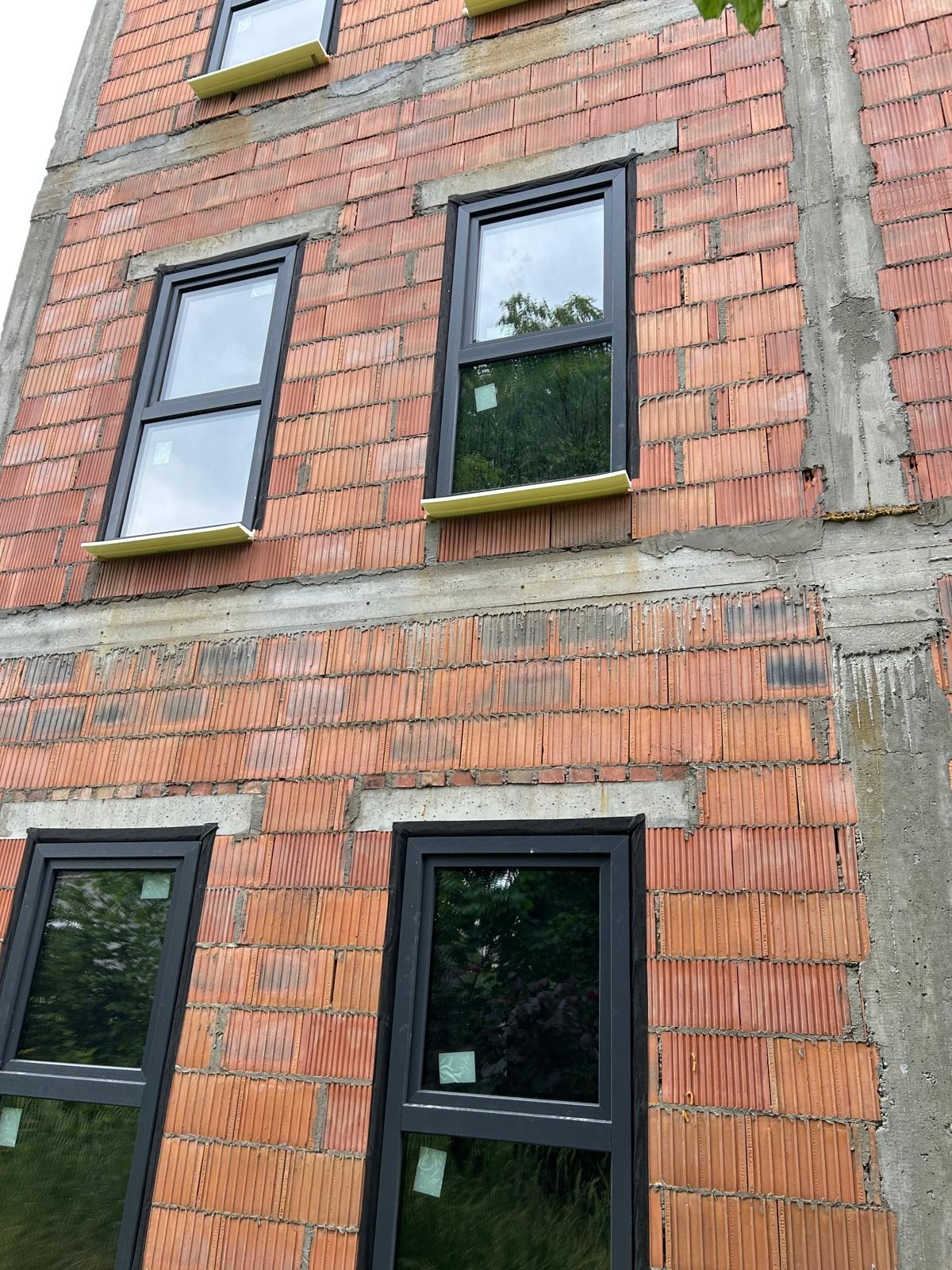 Montaż czarnych ram okiennych w surowej ścianie z czerwonej cegły dziurawki na budowie w Bytomiu, widoczne żółte podkładki pod oknami.