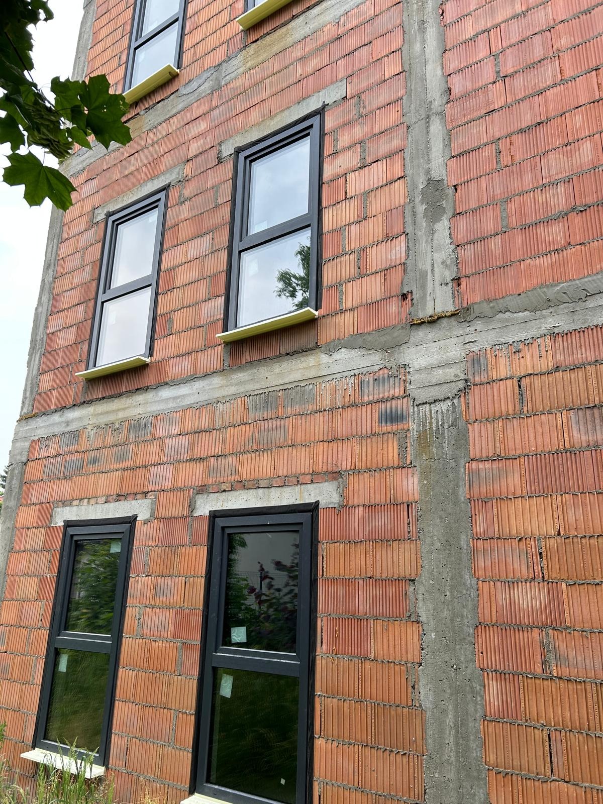 Surowa ściana z czerwonej cegły z czarnymi oknami, na których widać odbicie drzew. Widoczne nadproża i fragmenty konstrukcji betonowej. Okna częściowo zabezpieczone folią.