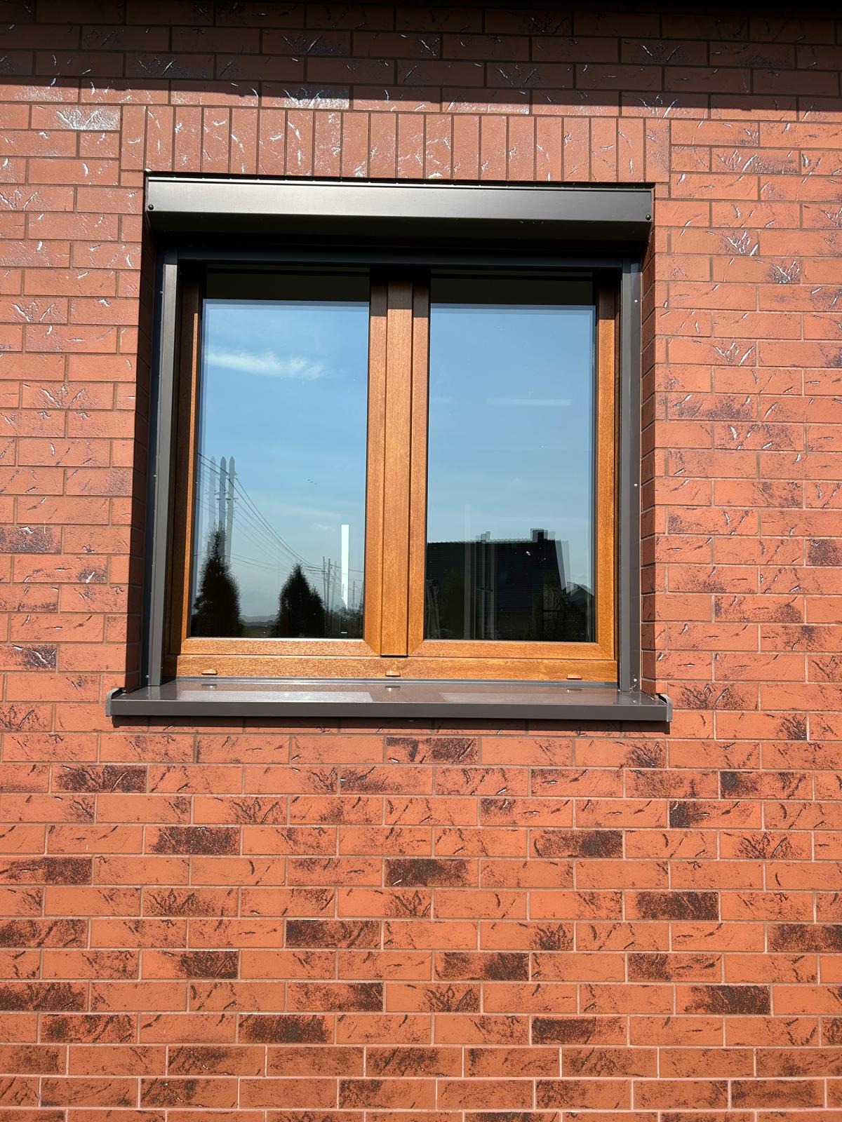 Dwuskrzydłowe okno z brązową ramą, zamontowane w elewacji z czerwonej cegły, z częściowo opuszczoną roletą w kolorze antracytowym.