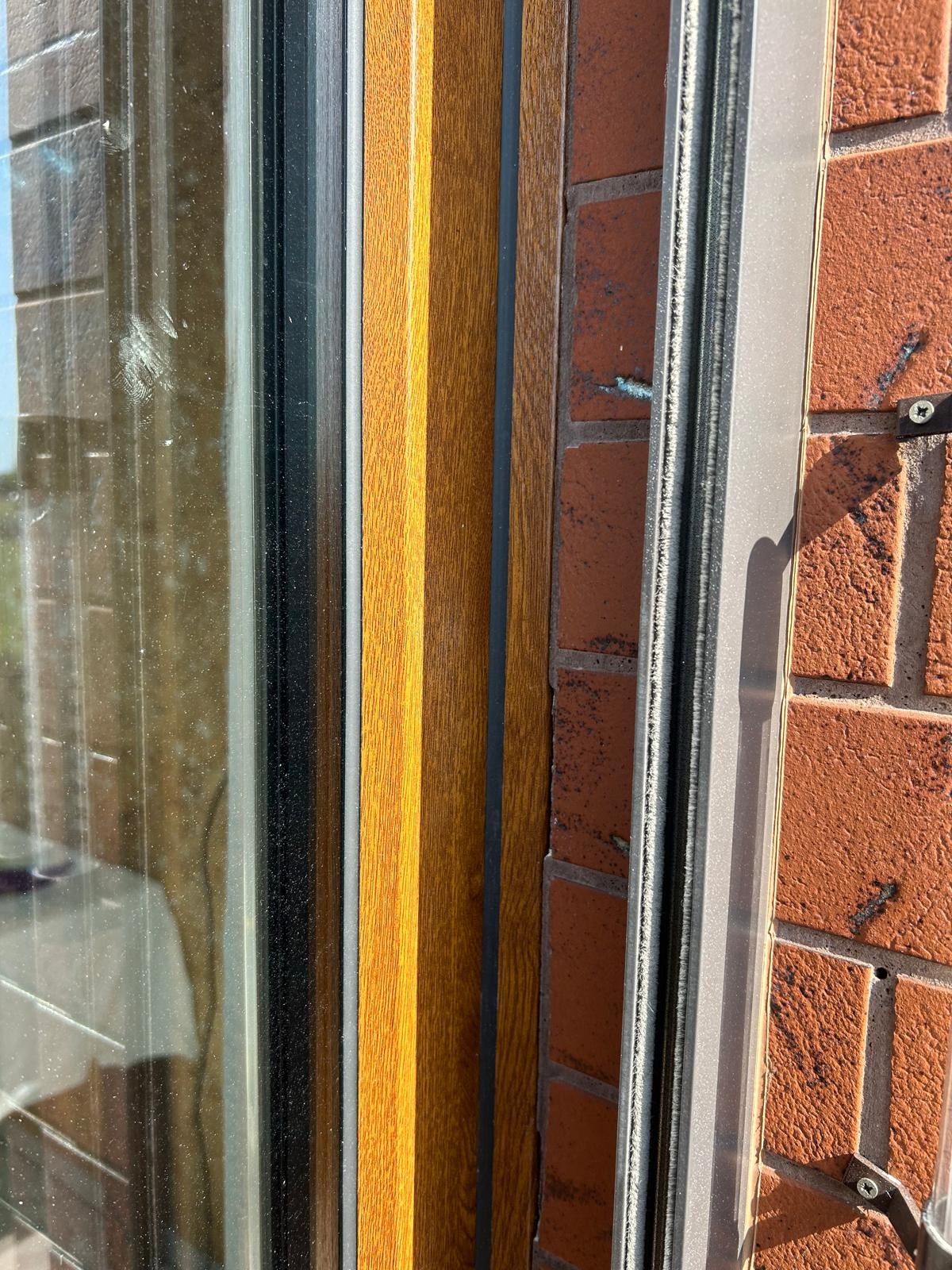 Fragment okna z drewnopodobną ramą i szybą, widoczna ceglana ściana z silikonem uszczelniającym, detale montażu.