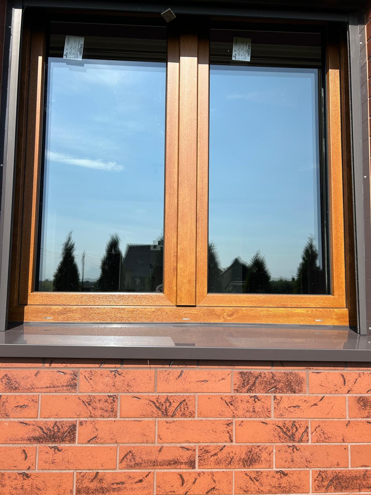 Dwuskrzydłowe okno z brązową ramą, widok z zewnątrz na ceglaną ścianę i odbicie nieba w szybach.