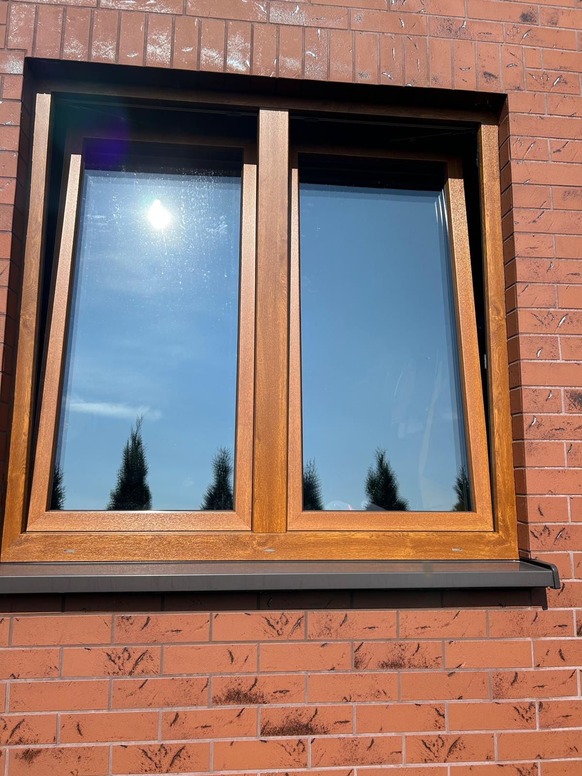 Drewniane okno PCV z widokiem na błękitne niebo i odbijające się w szybach drzewa iglaste, zamontowane w ścianie z czerwonej cegły.