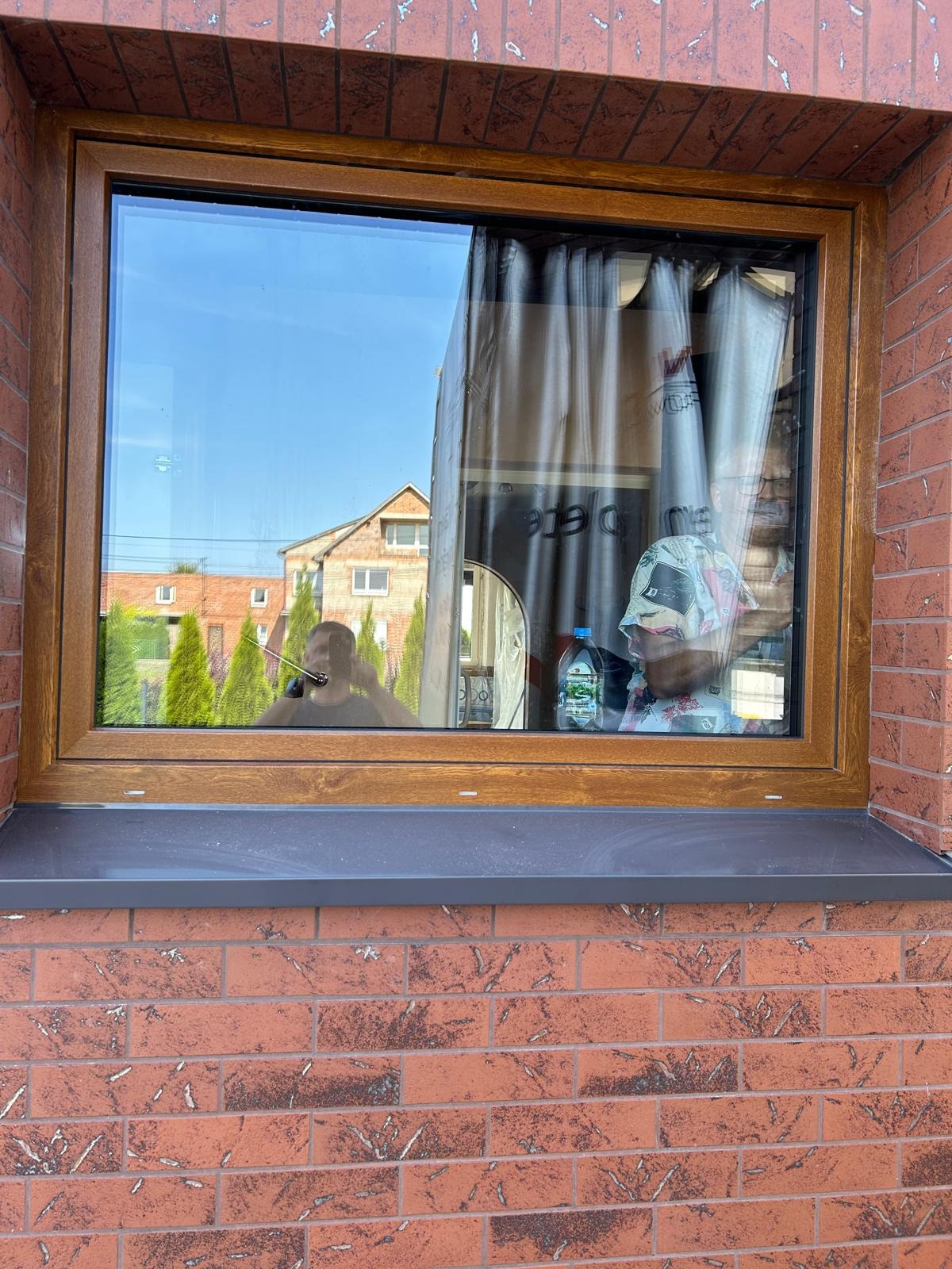 Zamontowane okno z brązową ramą w murze z czerwonej cegły, widoczne odbicie osoby robiącej zdjęcie i częściowo zasłonięte wnętrze folią ochronną i zasłonami.