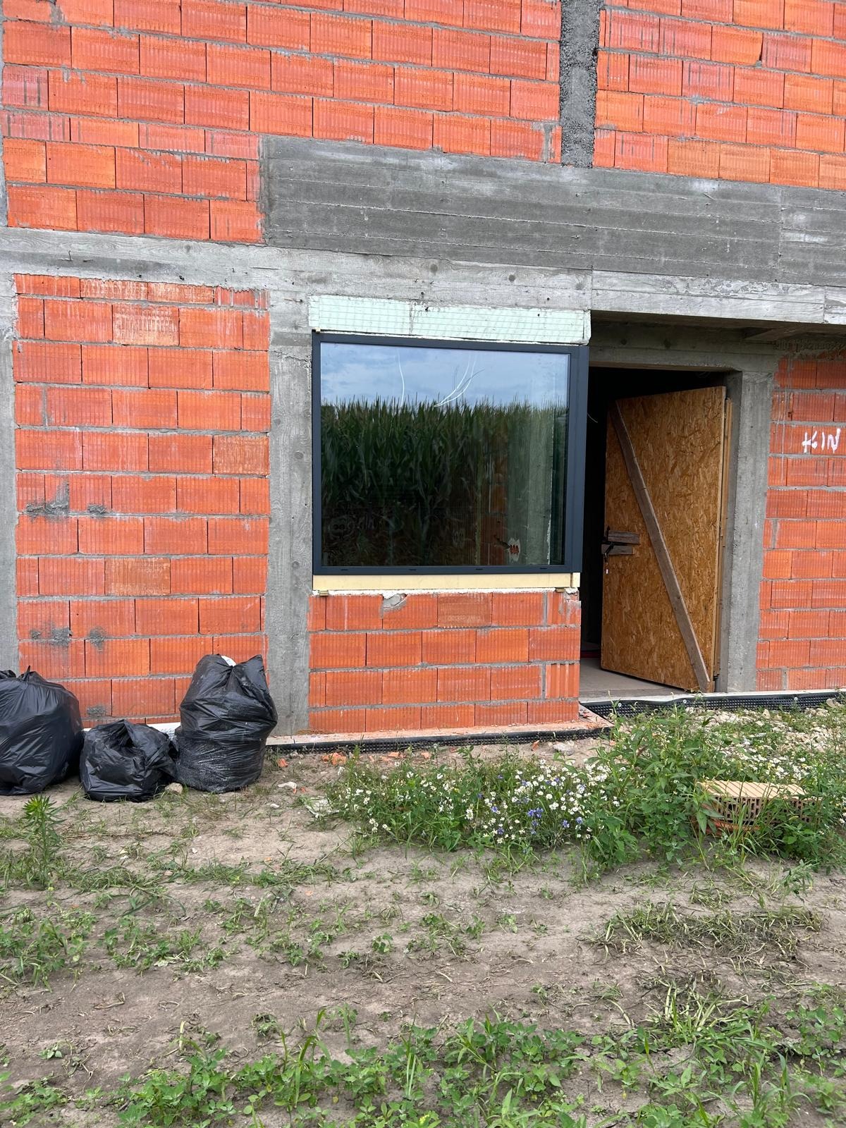 Surowy stan budynku w trakcie budowy z zamontowanym oknem z czarną ramą, widok na pole kukurydzy w odbiciu szyby, czarne worki na gruz i prowizoryczne drzwi.