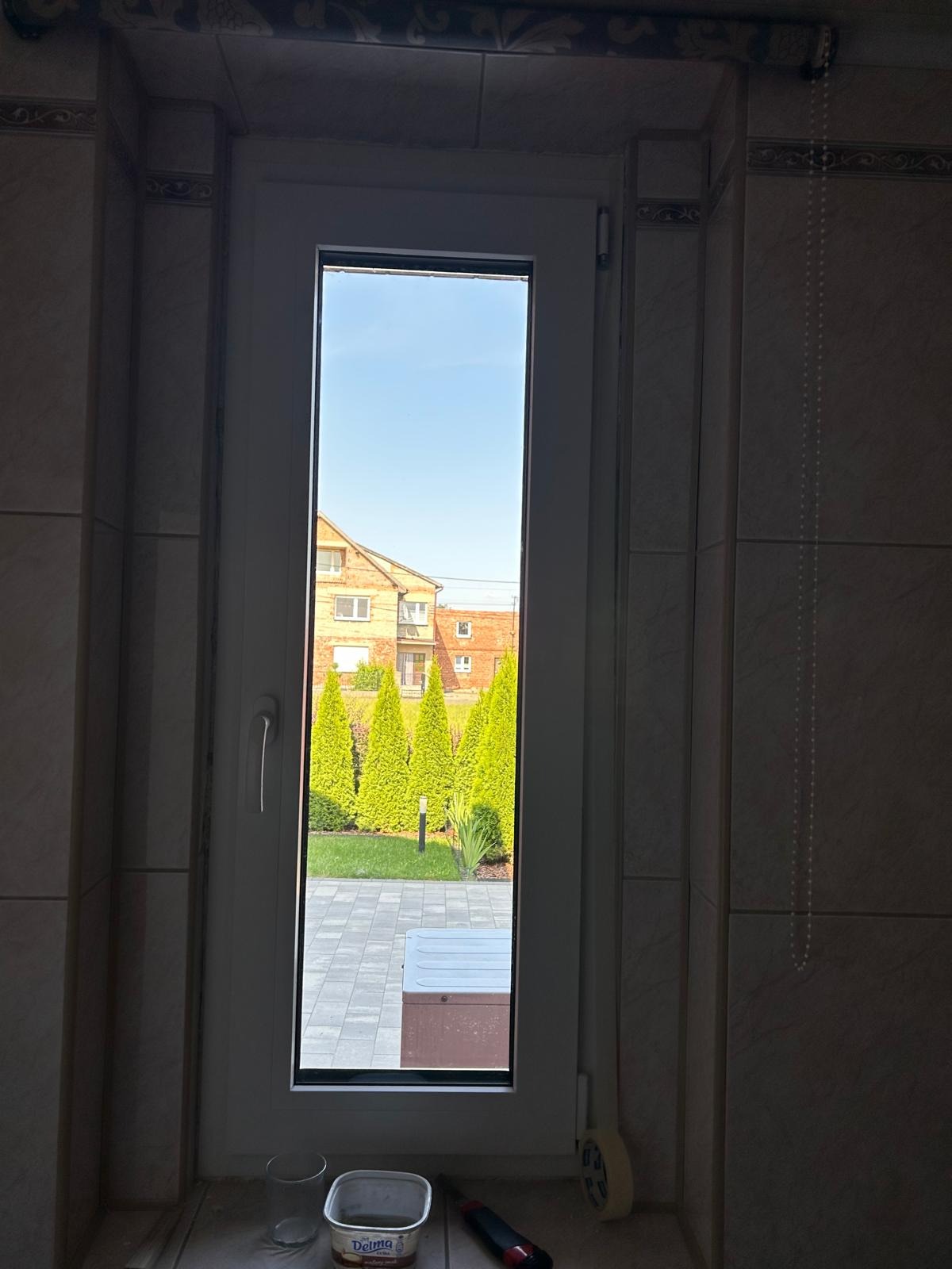 Wnętrze z widokiem na ogród przez pionowe okno w białej ramie, widoczne narzędzia na parapecie i fragment ściany z płytek.
