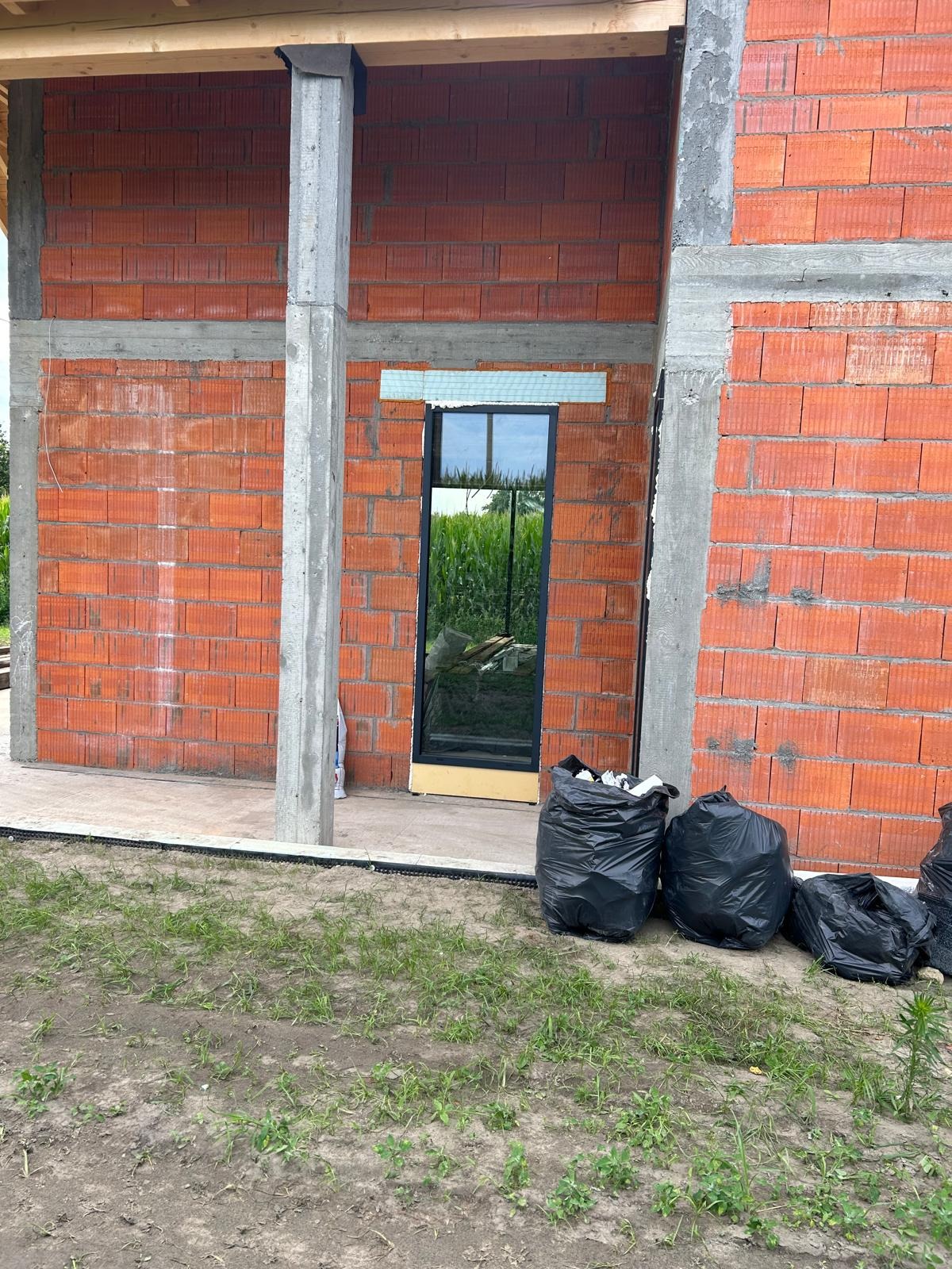 Surowy stan budowy: wstawione okno z czarną ramą w surowej ścianie z czerwonej cegły, widoczne worki z odpadami budowlanymi na pierwszym planie, tło z zielenią.