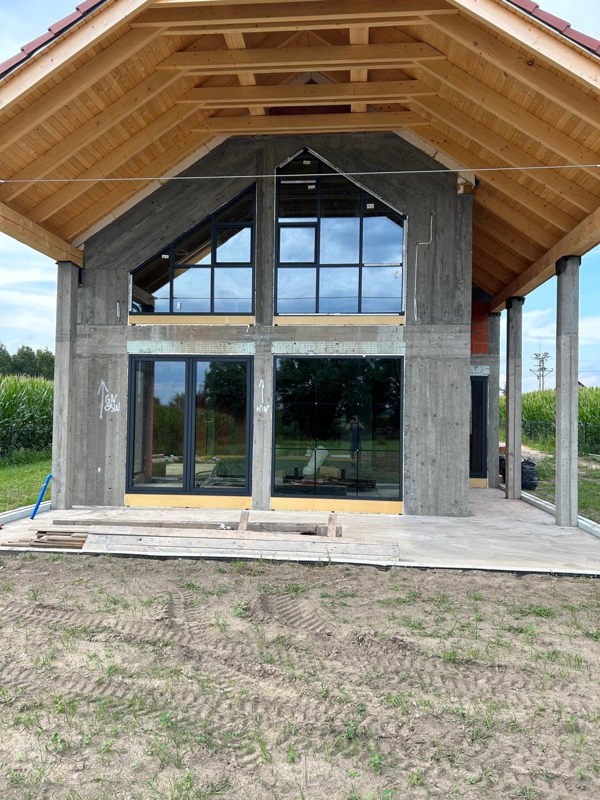 Surowy stan domu w trakcie budowy z zamontowanymi oknami o czarnych ramach, widoczne drewniane elementy konstrukcji dachu i surowe betonowe ściany.