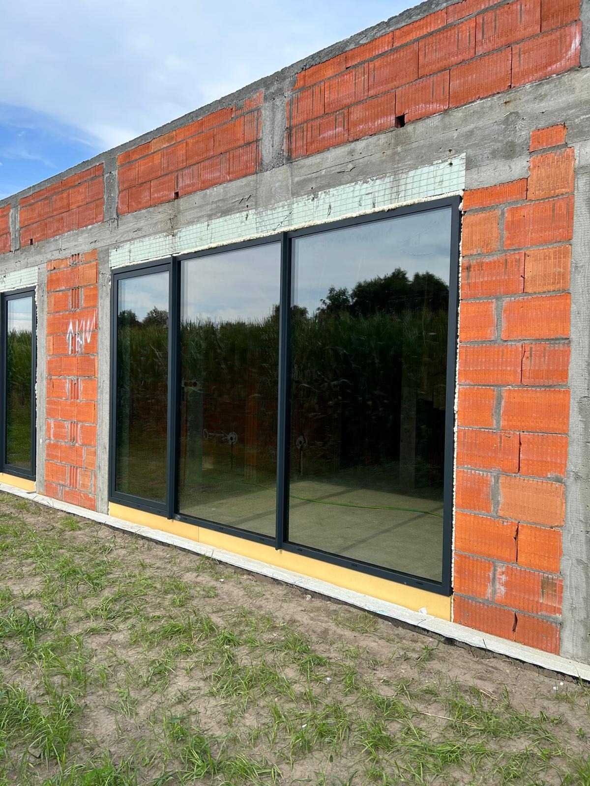 Czarne okna w surowym stanie budynku z czerwonej cegły, widoczne od zewnątrz na tle zieleni i nieba. Budynek w trakcie budowy, z widocznymi elementami konstrukcyjnymi i izolacyjnymi.