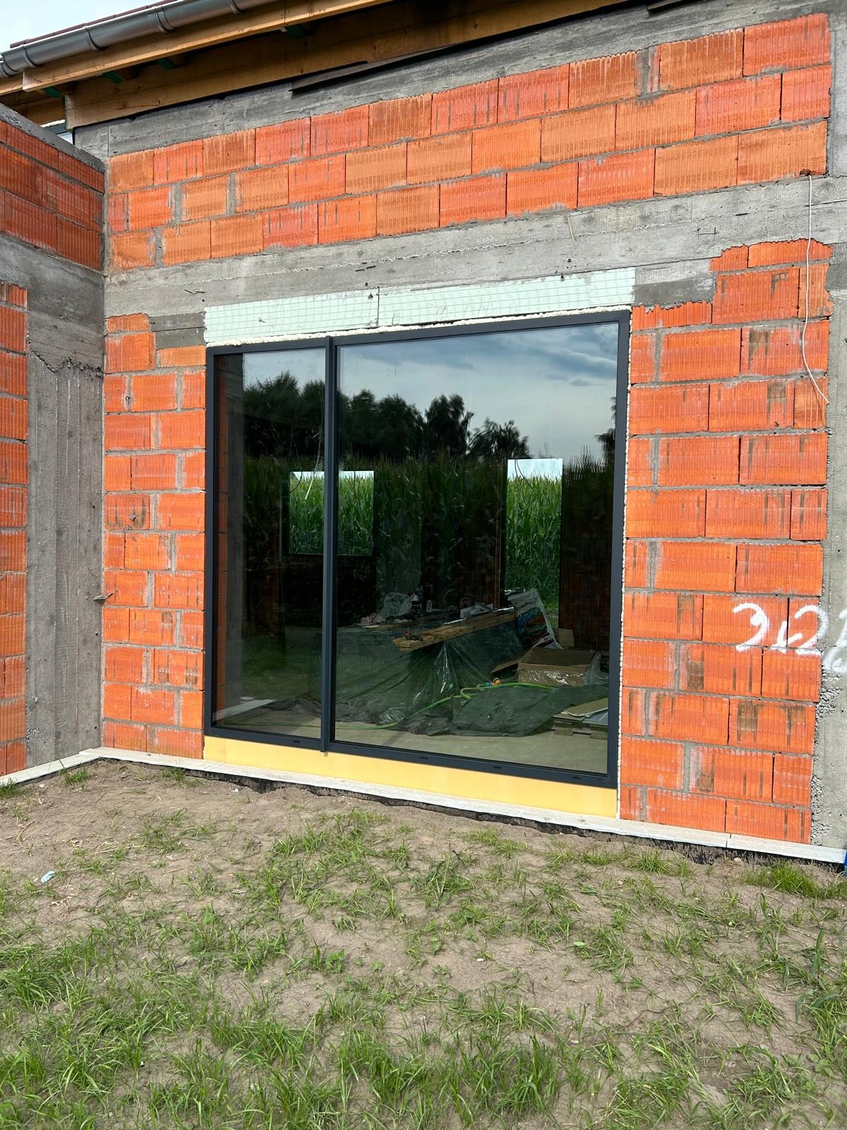 Montaż okna aluminiowego w surowym murze z czerwonej cegły, widoczne narzędzia i folia zabezpieczająca wewnątrz budynku, pole kukurydzy w odbiciu szyby.