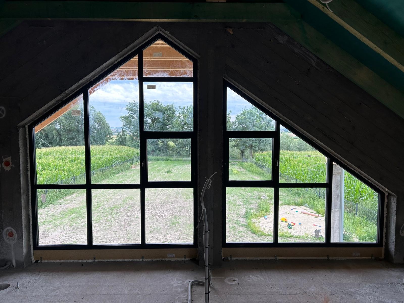 Dwa okna o nietypowym, trójkątnym kształcie w surowym, betonowym wnętrzu, z widokiem na zielone pole kukurydzy i ogród z piaskownicą.