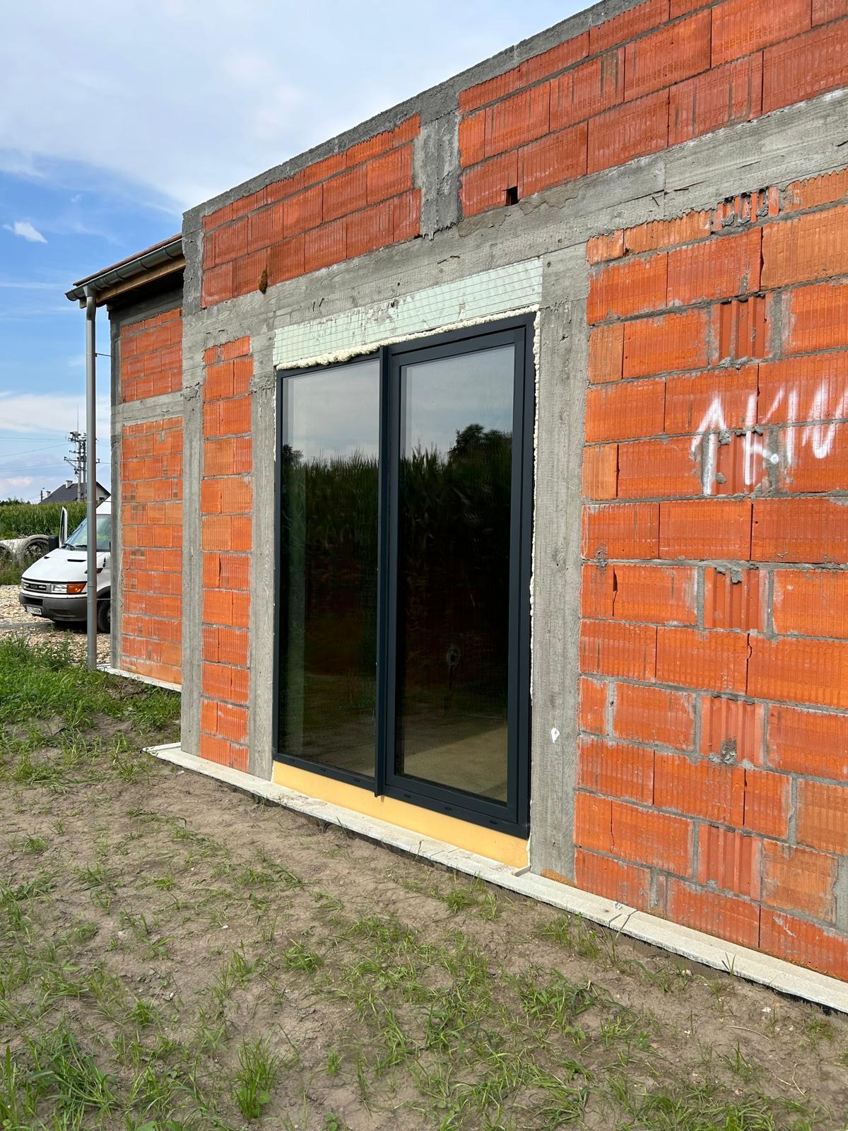 Surowe ściany z czerwonej cegły z zamontowanym przesuwnym oknem z ciemną ramą, widoczne elementy izolacji i fundamentu, w tle pole kukurydzy i biały van.