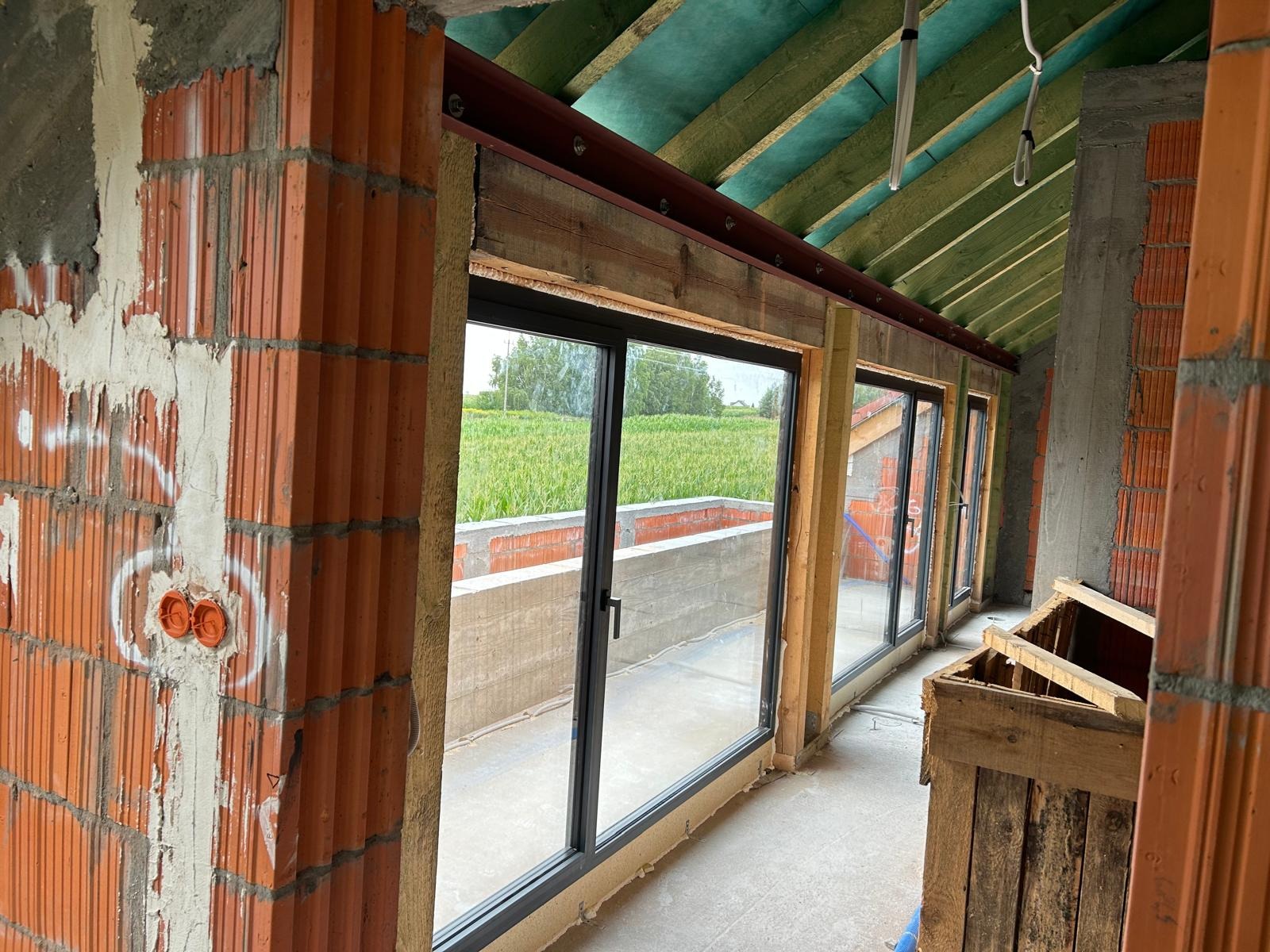 Wnętrze budynku w trakcie budowy z zamontowanymi oknami z czarnymi ramami; widok na pole za oknami, surowy stan wykończenia ścian z cegły i betonu, drewniana konstrukcja dachu.